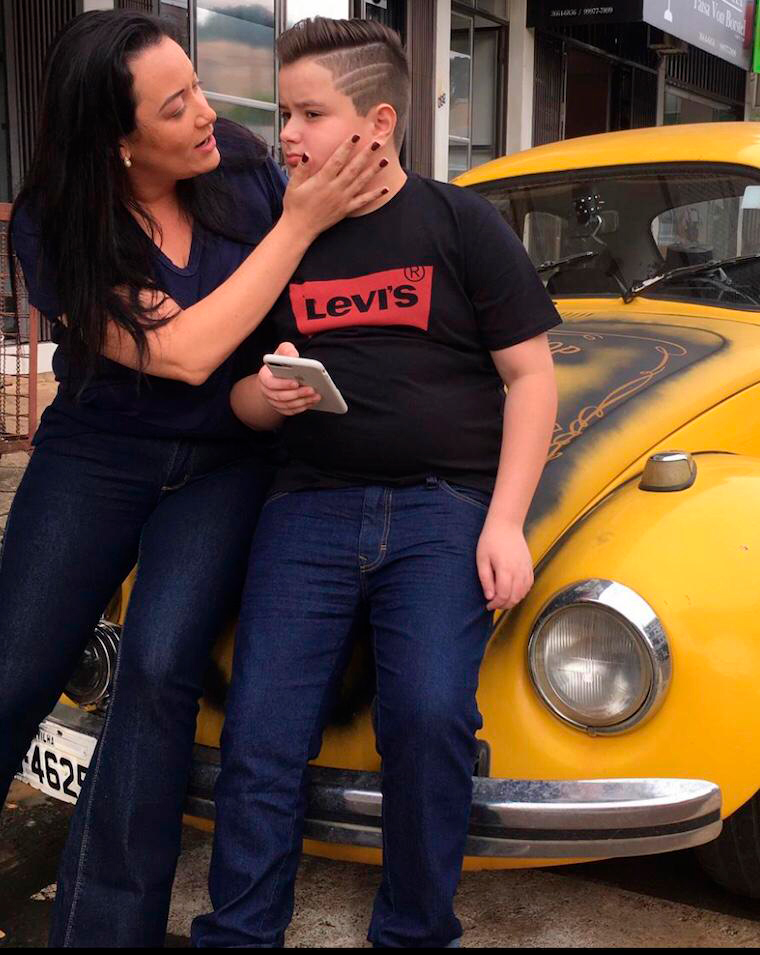 Andrea com o filho João Pedro. Estão de pé, encostados em um carro (Fusca) amarelo. Eles vestem calça jeans e camiseta de cor escura. A mãe está olhando para o filho e com a mão no rosto dele. O garoto segura um celular.