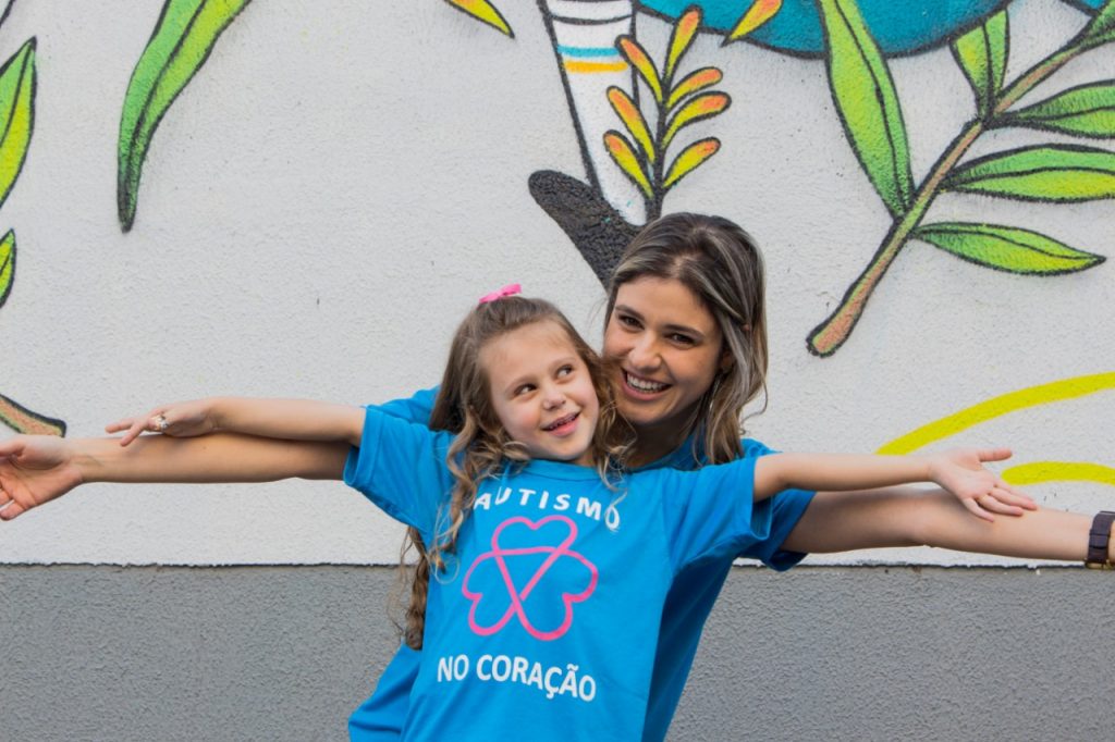 Alice e Adriana, de braços abertos, entrelaçados, sorriem e usam camiseta azul com o texto Autismo no Coração.