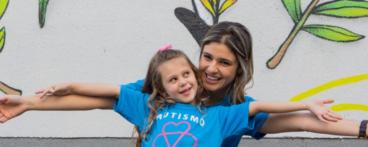 Alice e Adriana, de braços abertos, entrelaçados, sorriem e usam camiseta azul com o texto Autismo no Coração.