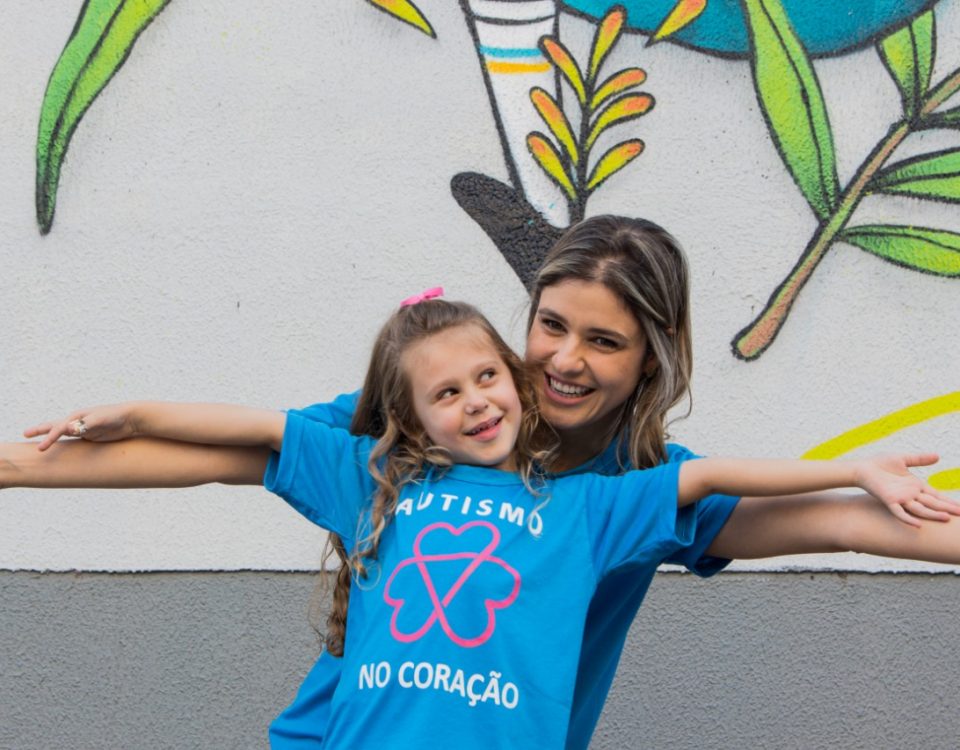 Alice e Adriana, de braços abertos, entrelaçados, sorriem e usam camiseta azul com o texto Autismo no Coração.