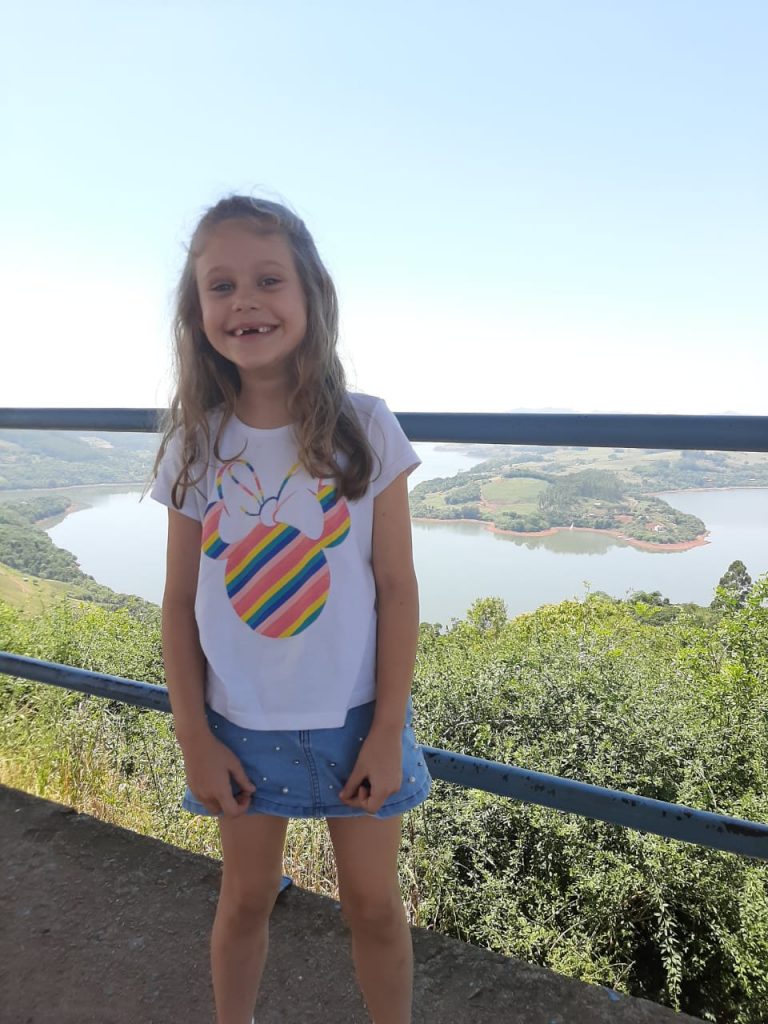 Alice sorrindo, com bela paisagem ao fundo (lago e árvores). A menina é loira, tem cabelos longos e ondulados. Veste saia jeans e camiseta branca com contorno colorido da personagem Minnie.