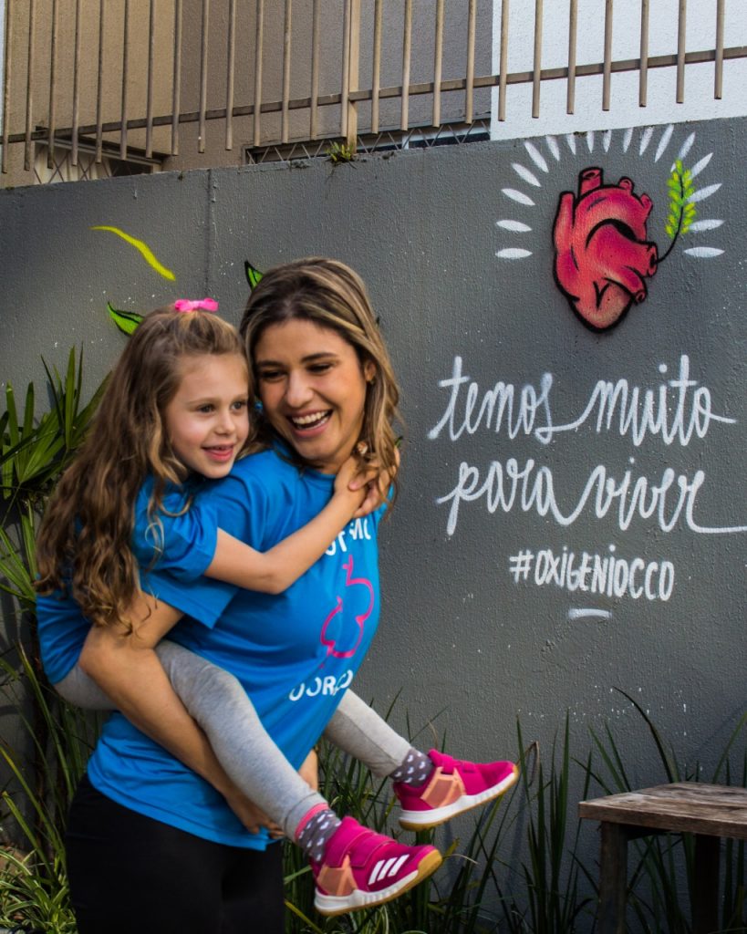 Alice e Adriana brincam e sorriem, Adriana carrega a filha nas costas. Ambas vestem camiseta azul