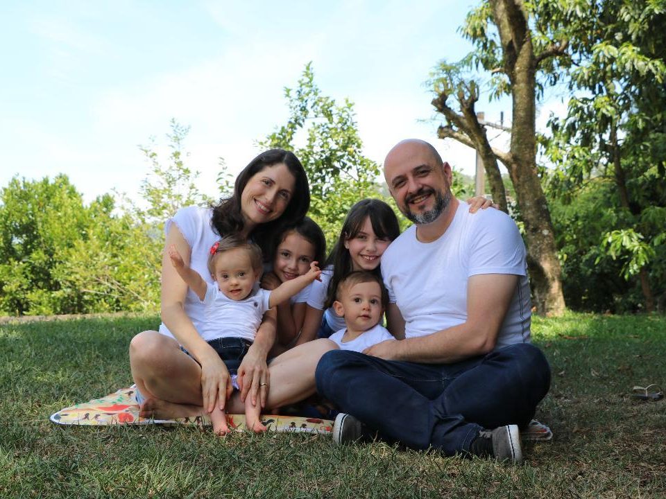 A família está sentada em um gramado, rodeada por árvores. Todos usam camiseta branca e sorriem.