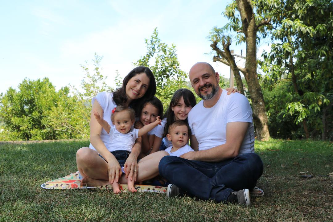 A família está sentada em um gramado, rodeada por árvores. Todos usam camiseta branca e sorriem.