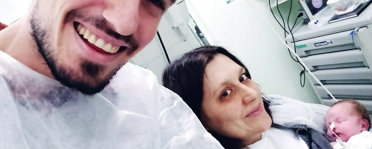 Jean e Eliandra vestem avental hospitalar branco. Eliandra segura o bebê no colo. Os pais sorriem, o filho dorme e usa sonda no nariz.