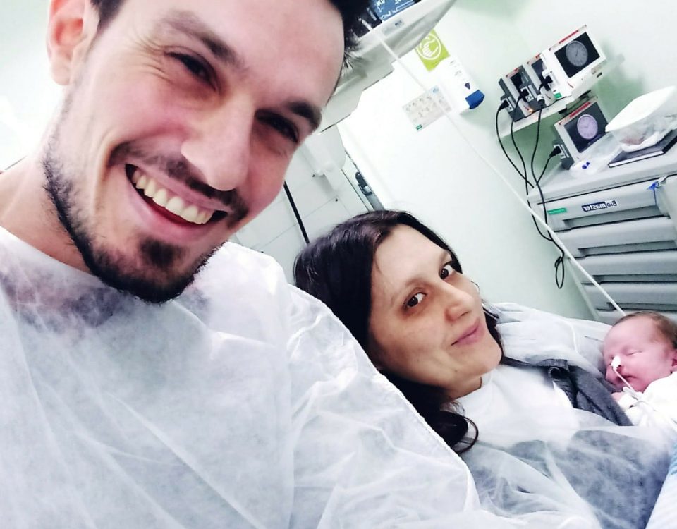 Jean e Eliandra vestem avental hospitalar branco. Eliandra segura o bebê no colo. Os pais sorriem, o filho dorme e usa sonda no nariz.