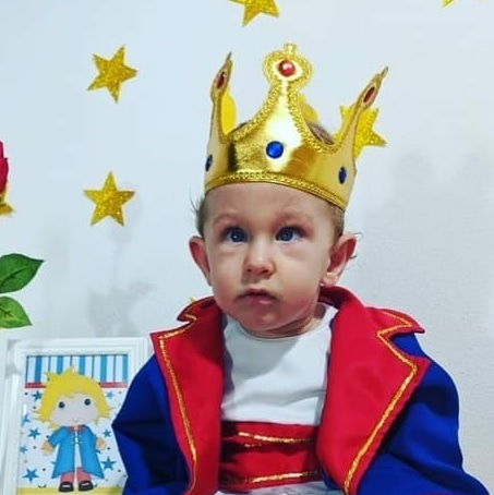 Giordano, menino loiro de olhos azuis, sentado, vestindo trajes de rei (capa e coroa).