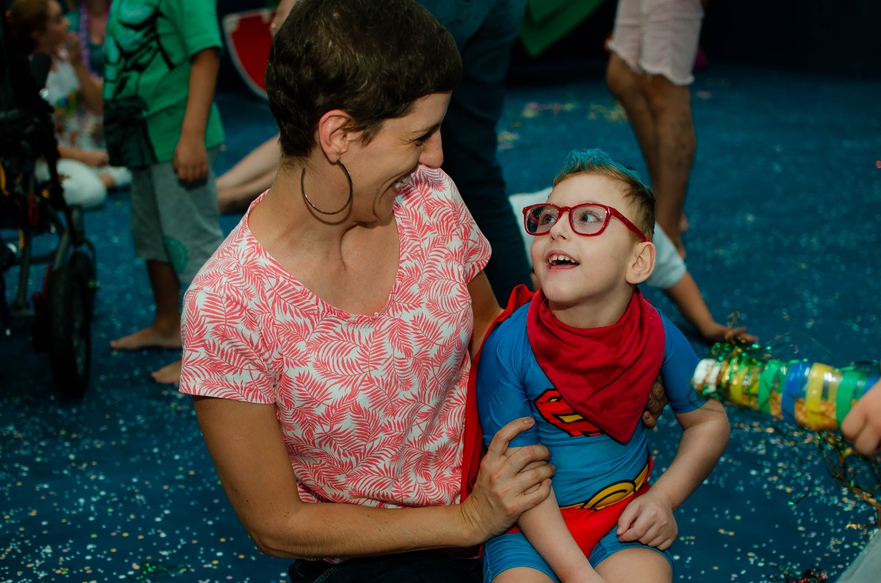 Fernanda e Caio em festa de carnaval, sentados no chão. Eles se olham com amor e alegria. Fernanda está com o cabelo bem curto e brinco grande (argola). Caio usa fantasia do Super Homem e óculos com armação vermelha.