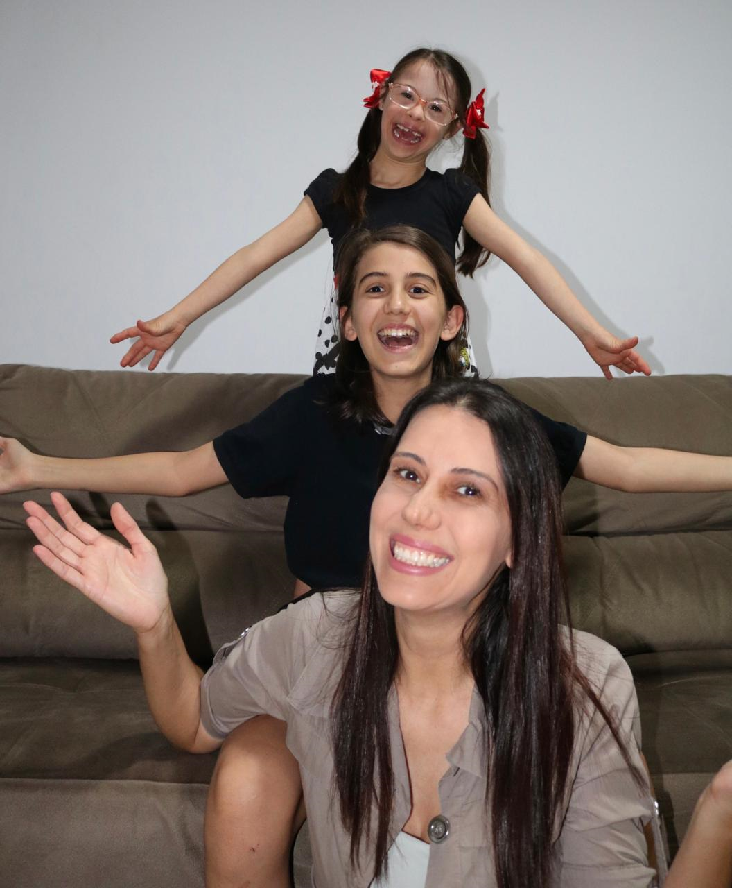 Juliana com os filhos Luís Gustavo, de 12 anos, e Ana Luísa, de 8, formando um totem, usando o sofá como apoio. Os três estão de bracos abertos e sorriem.