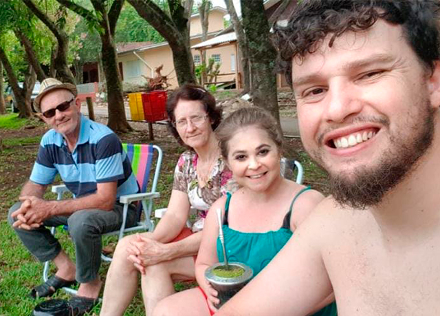 Gisely, o marido e os pais estão sentados em cadeiras de praia, em uma praça, tomando chimarrão.